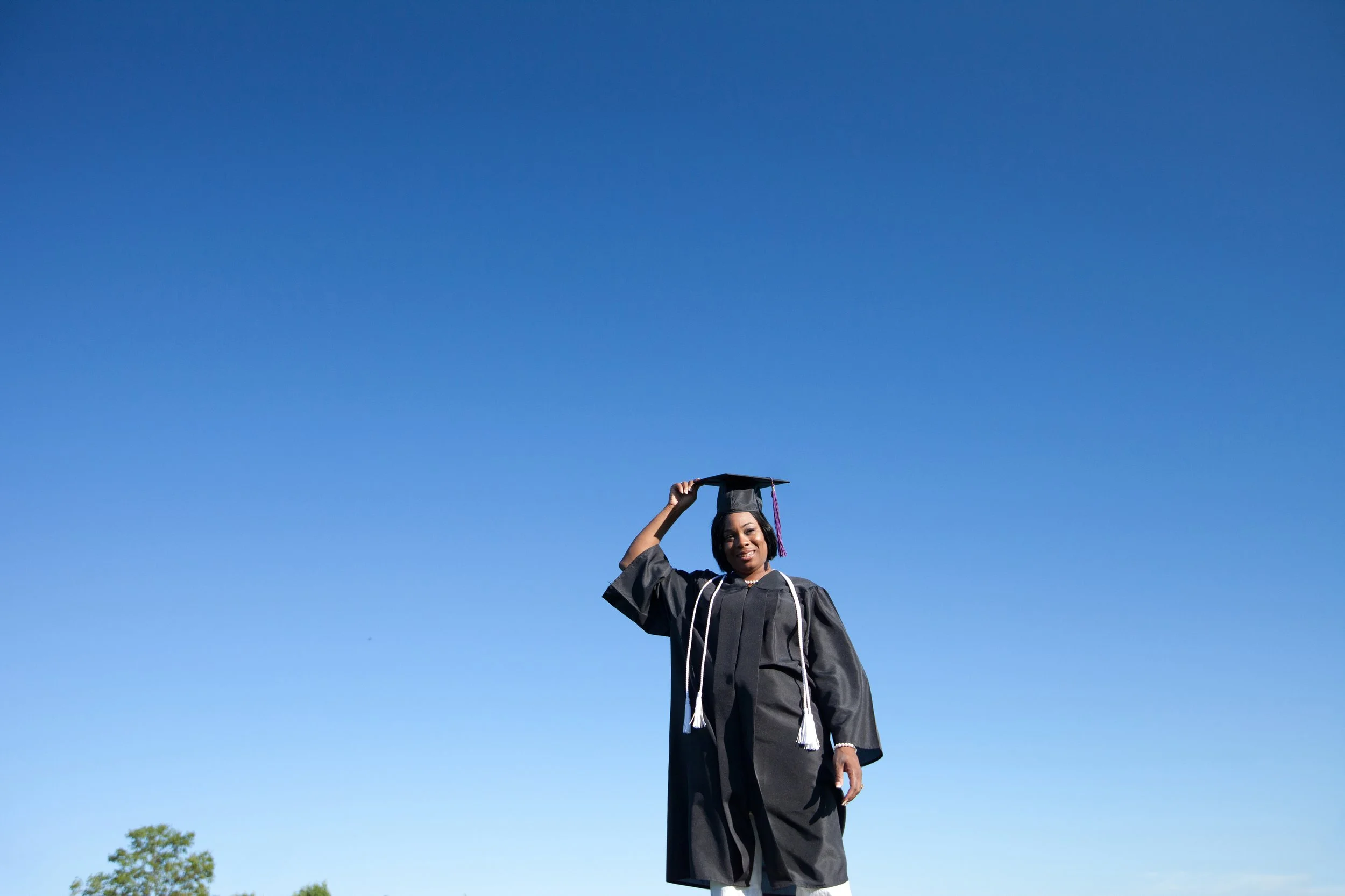 A woman in graduation cap and gown standing outdoors against a clear blue sky, smiling, with trees in the background.