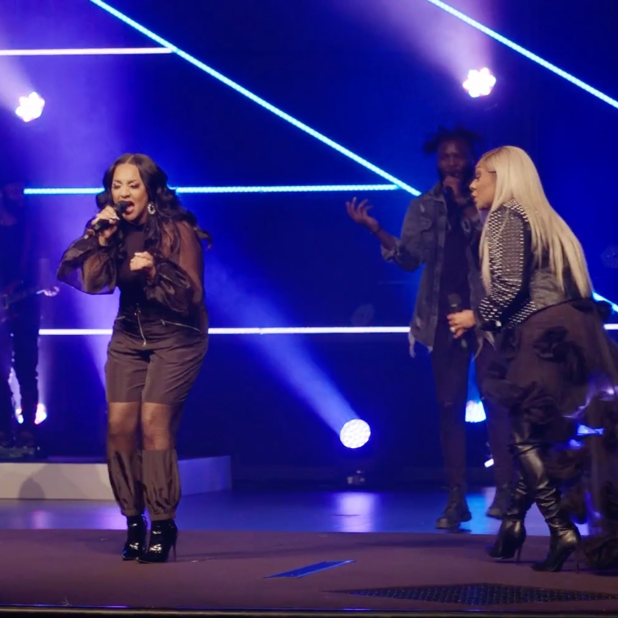 A female singer performing on stage with two people behind her, colorful stage lights and blue neon lines in the background.