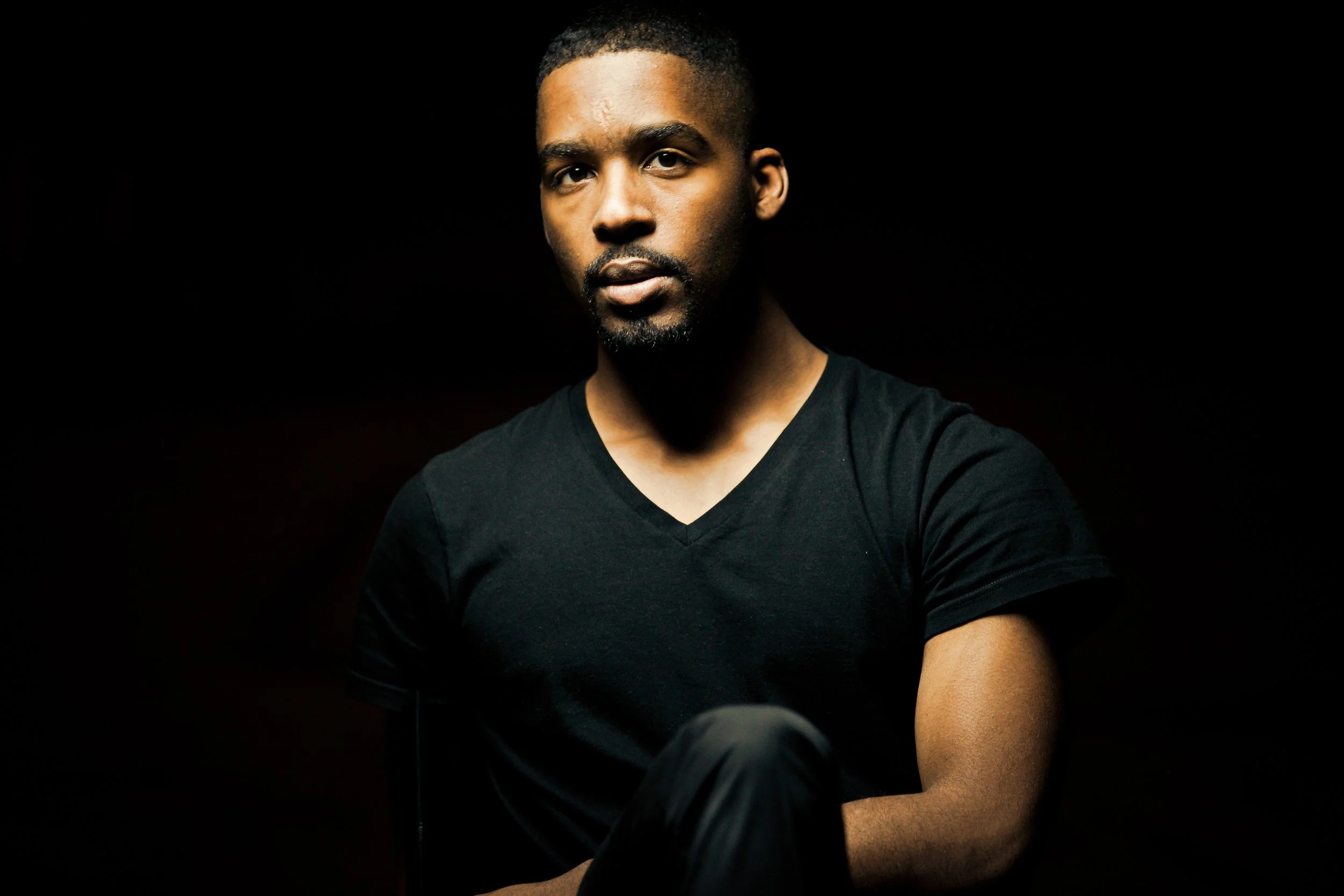Portrait of a man with short hair, beard, wearing a black V-neck shirt, against a dark background.