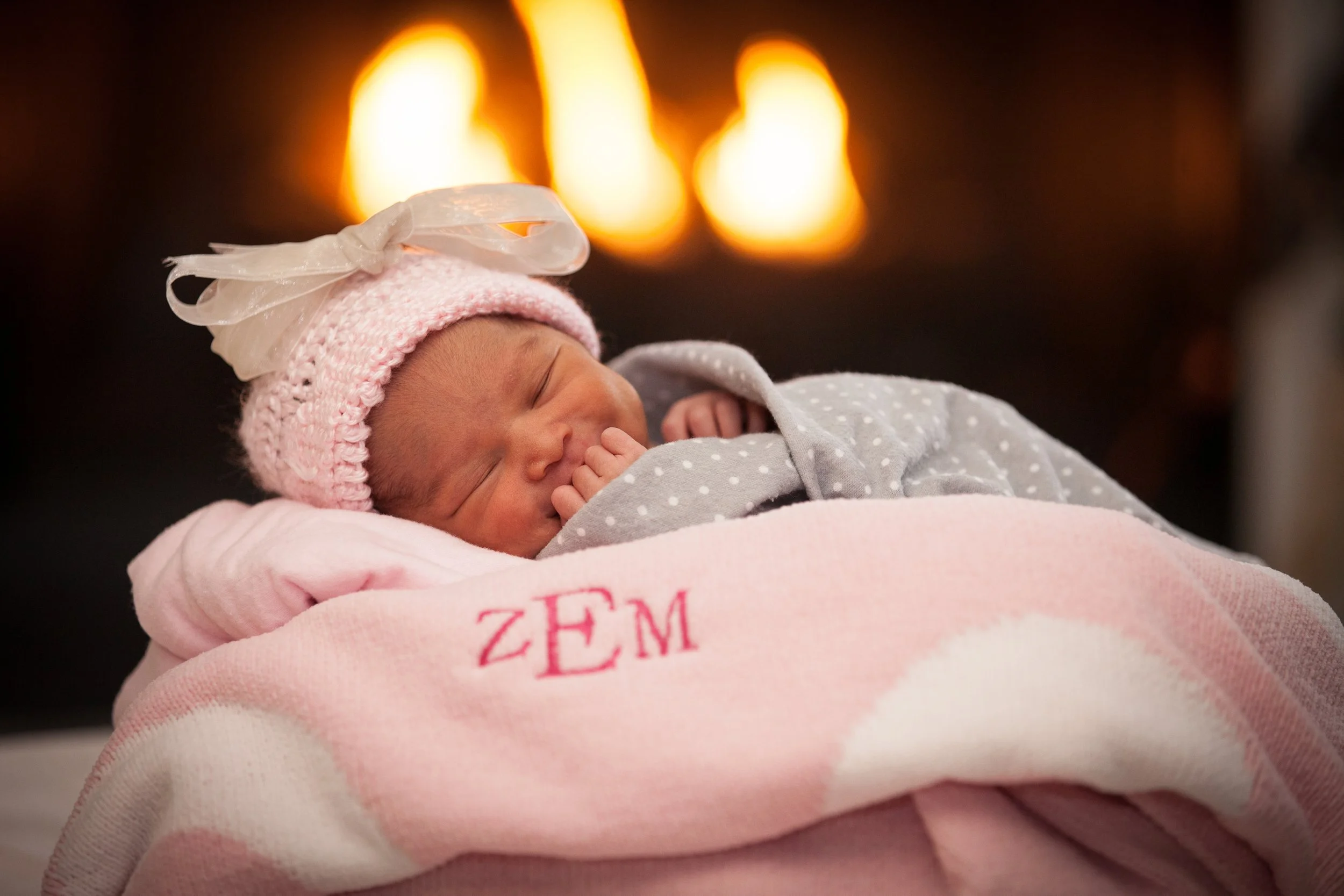 A newborn baby sleeping on a pink blanket with initials 'ZEM', wearing a pink knit hat with a white ribbon bow and a gray and white polka dot outfit, with a fireplace glowing in the background.