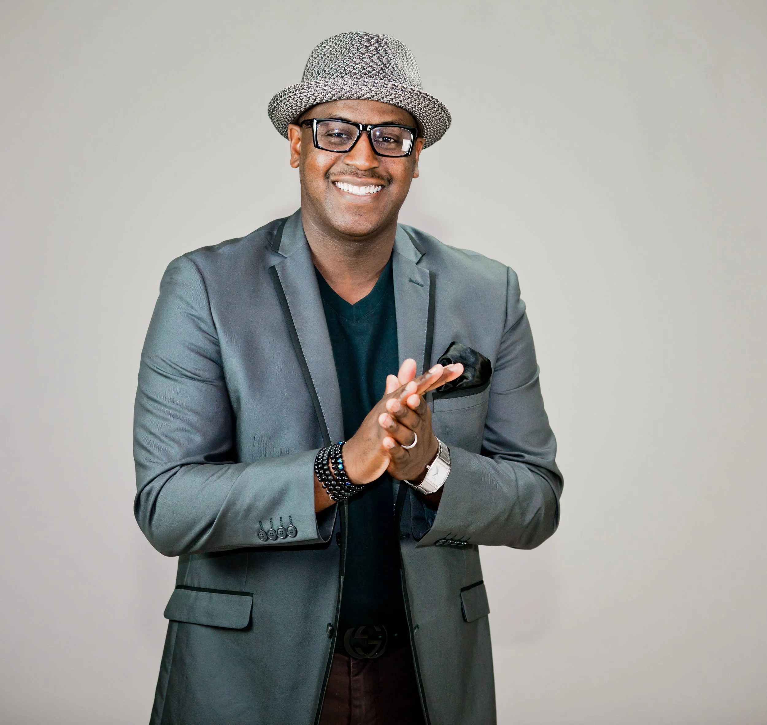 Smiling man wearing glasses, a gray blazer, black t-shirt, a matching hat, and accessories, posing with hands together against a plain background.