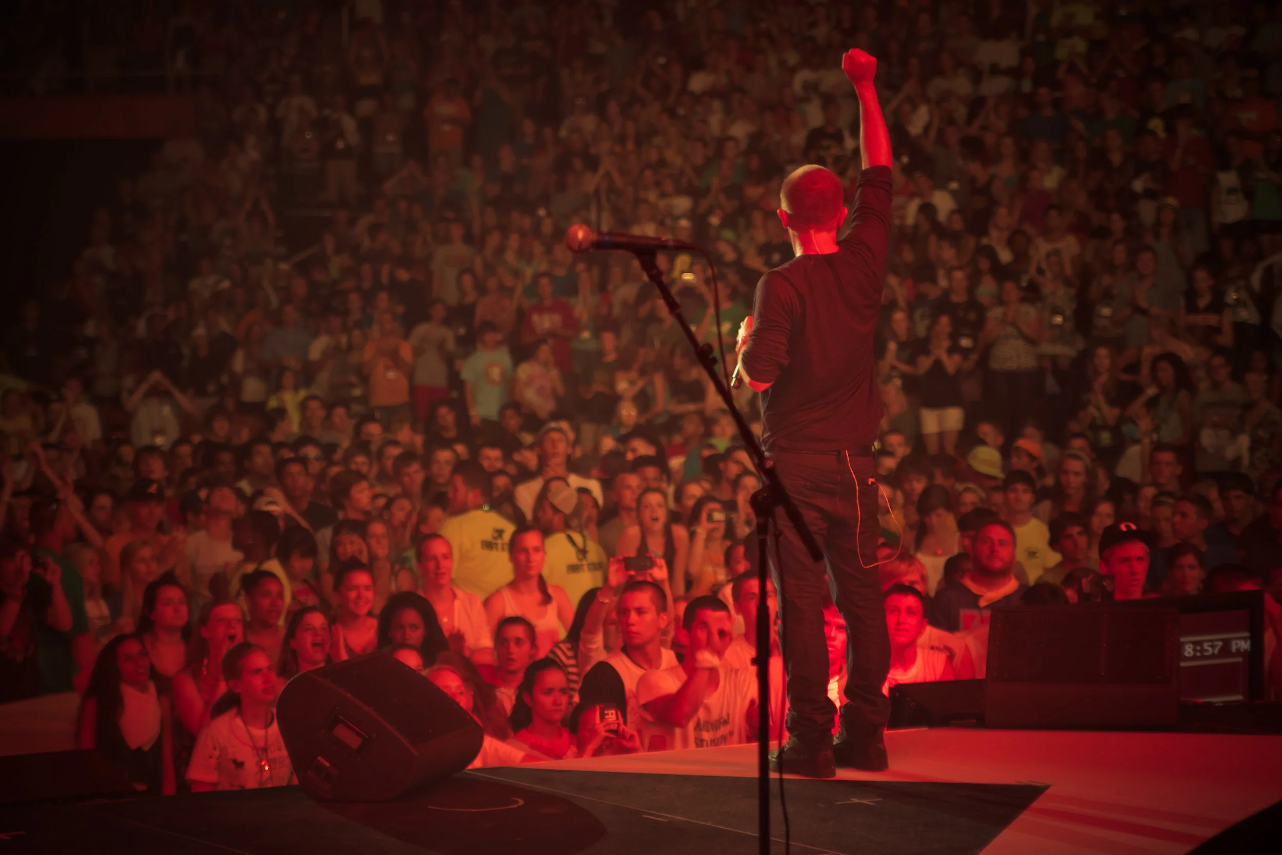 Performer on stage with arm raised facing an excited audience in a concert hall.