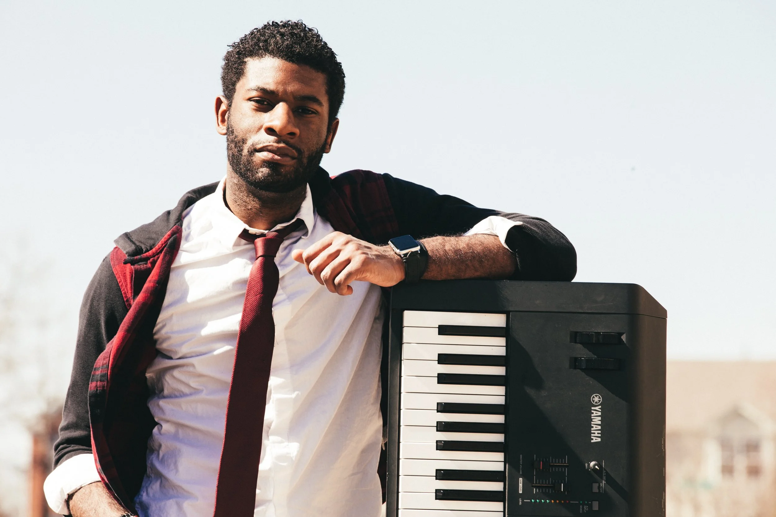 A man with dark hair and a beard leaning on a Yamaha keyboard outdoors during daytime, wearing a white shirt, red tie, dark jacket, and a smartwatch.
