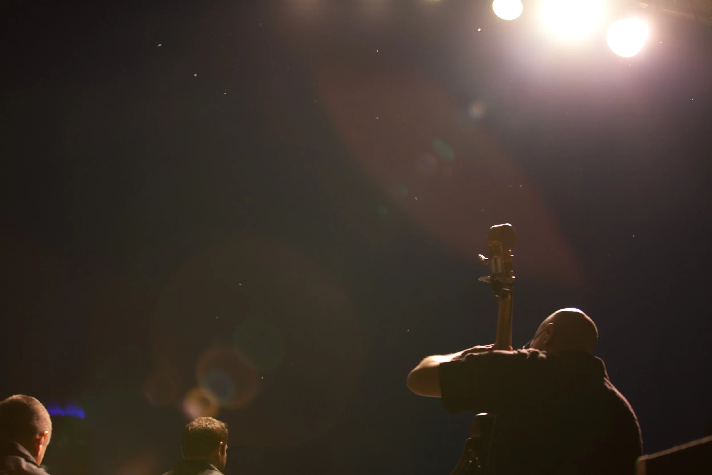 A person playing a guitar on stage under bright lights at night, with a dark sky in the background and other people visible in the foreground.