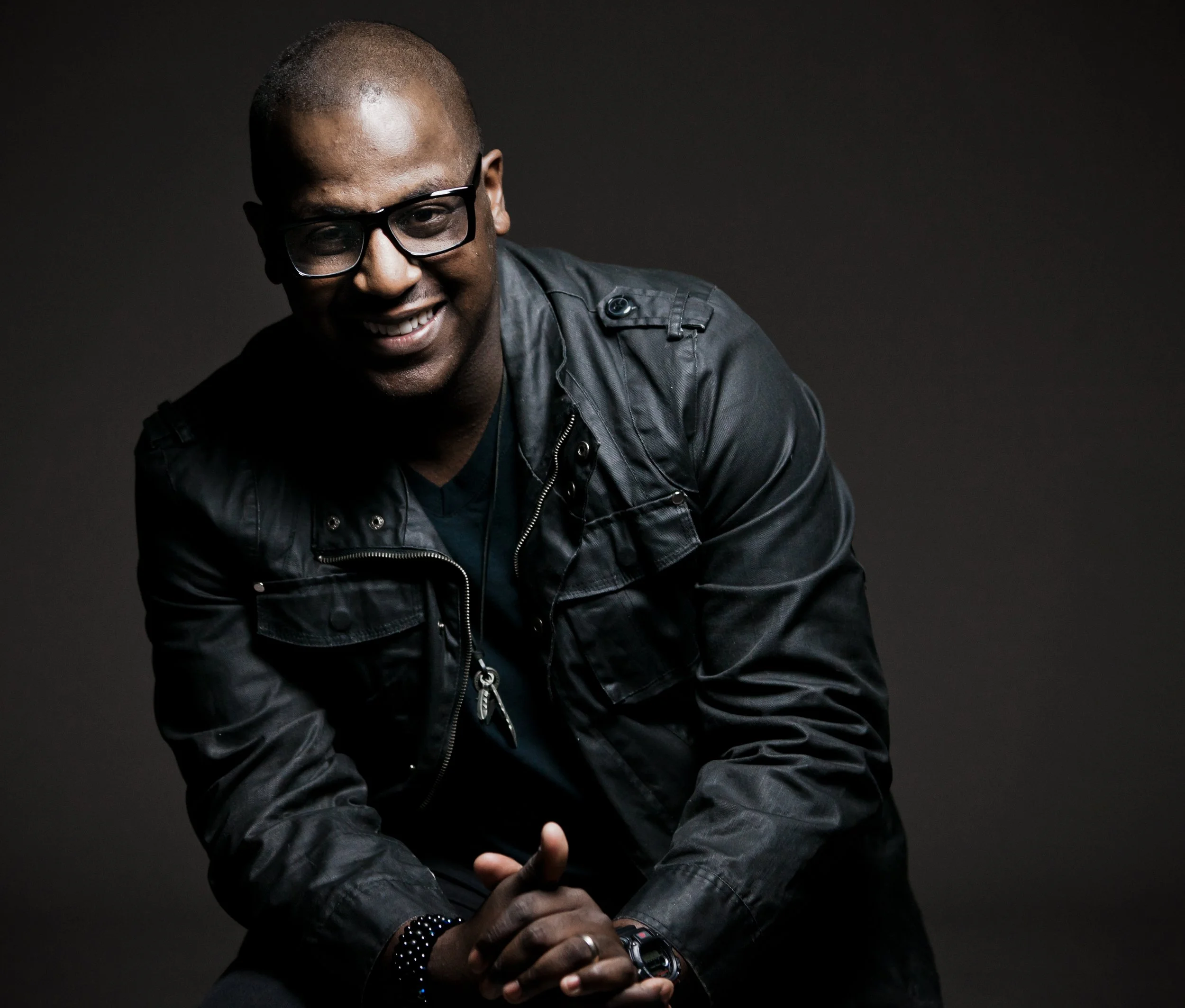 A man with glasses, smiling, wearing a black leather jacket, seated against a dark background.