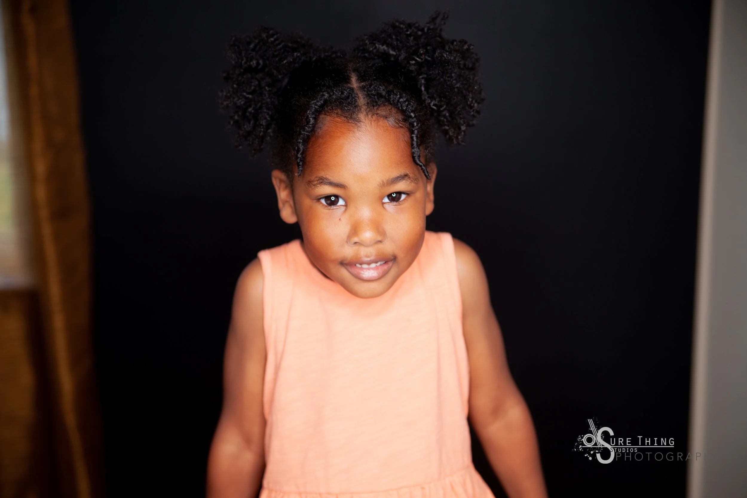 A young girl with curly hair styled in two puffs wearing a peach-colored dress standing in front of a black backdrop.