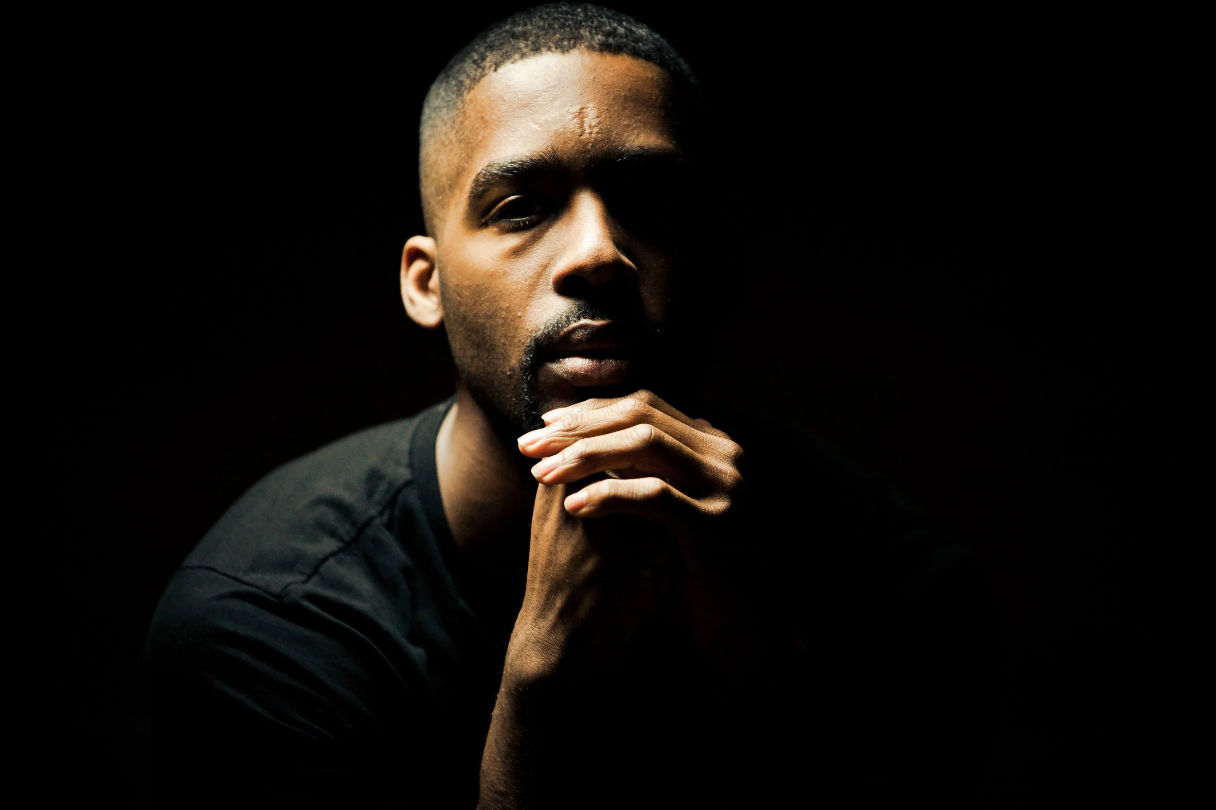 A man with a contemplative expression, resting his chin on his clasped hands, against a dark background.