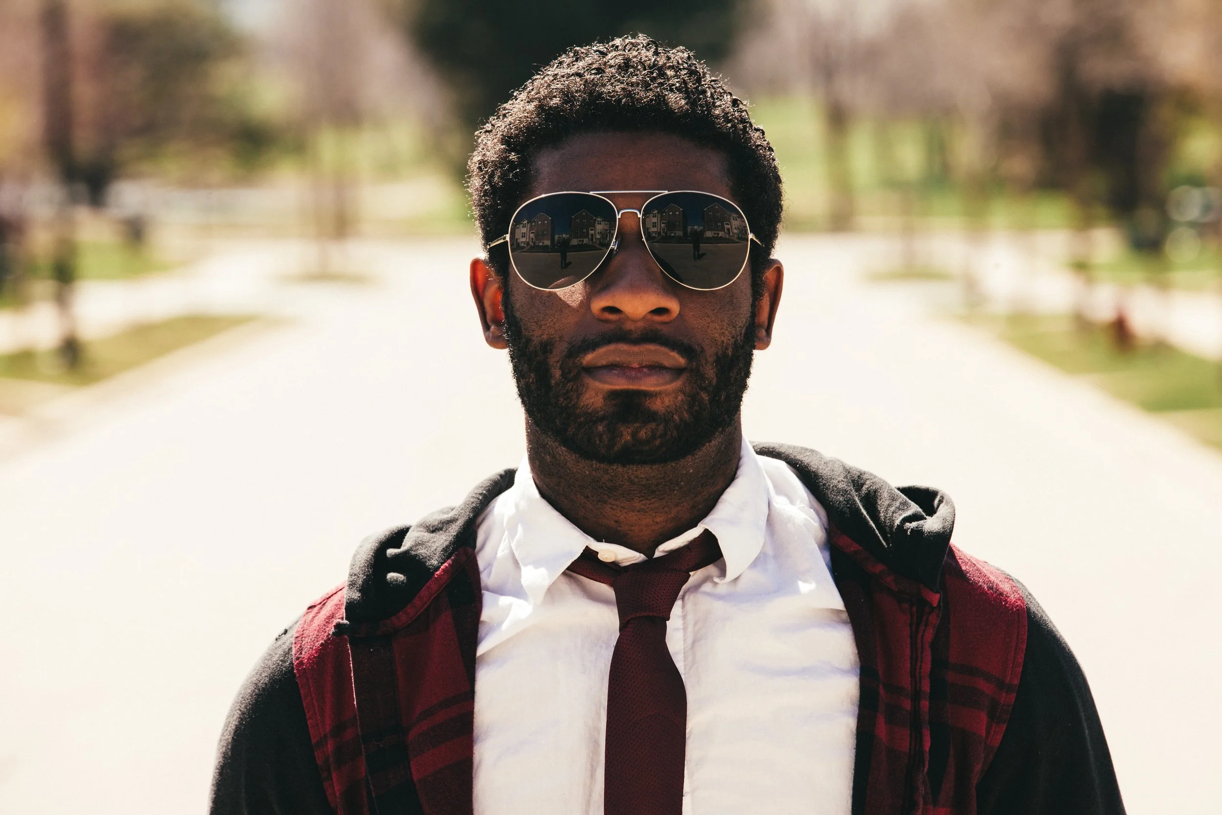 A man with curly hair and a beard wearing sunglasses, a white shirt, a dark tie, and a jacket, standing outdoors on a sunny day.
