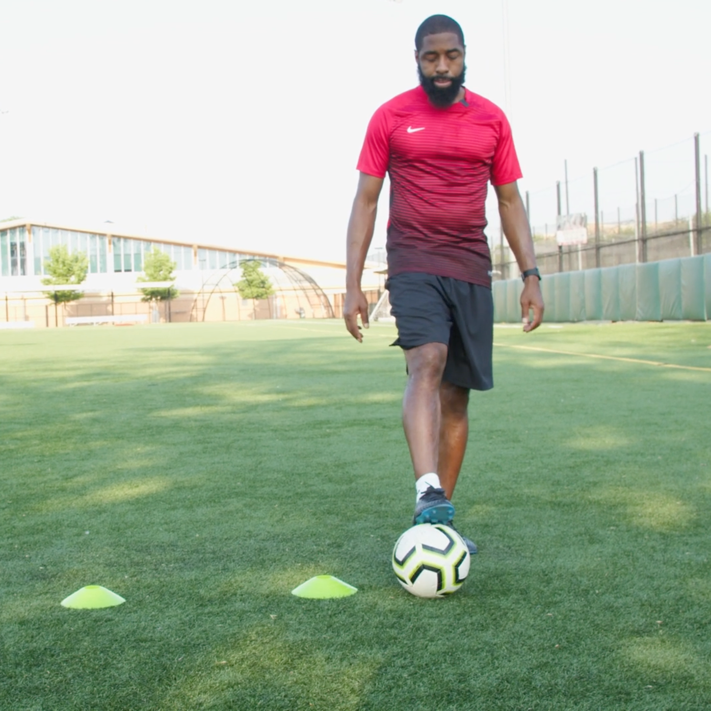 A man practicing soccer on a green field, walking with a soccer ball near his foot past yellow training cones, during daytime.