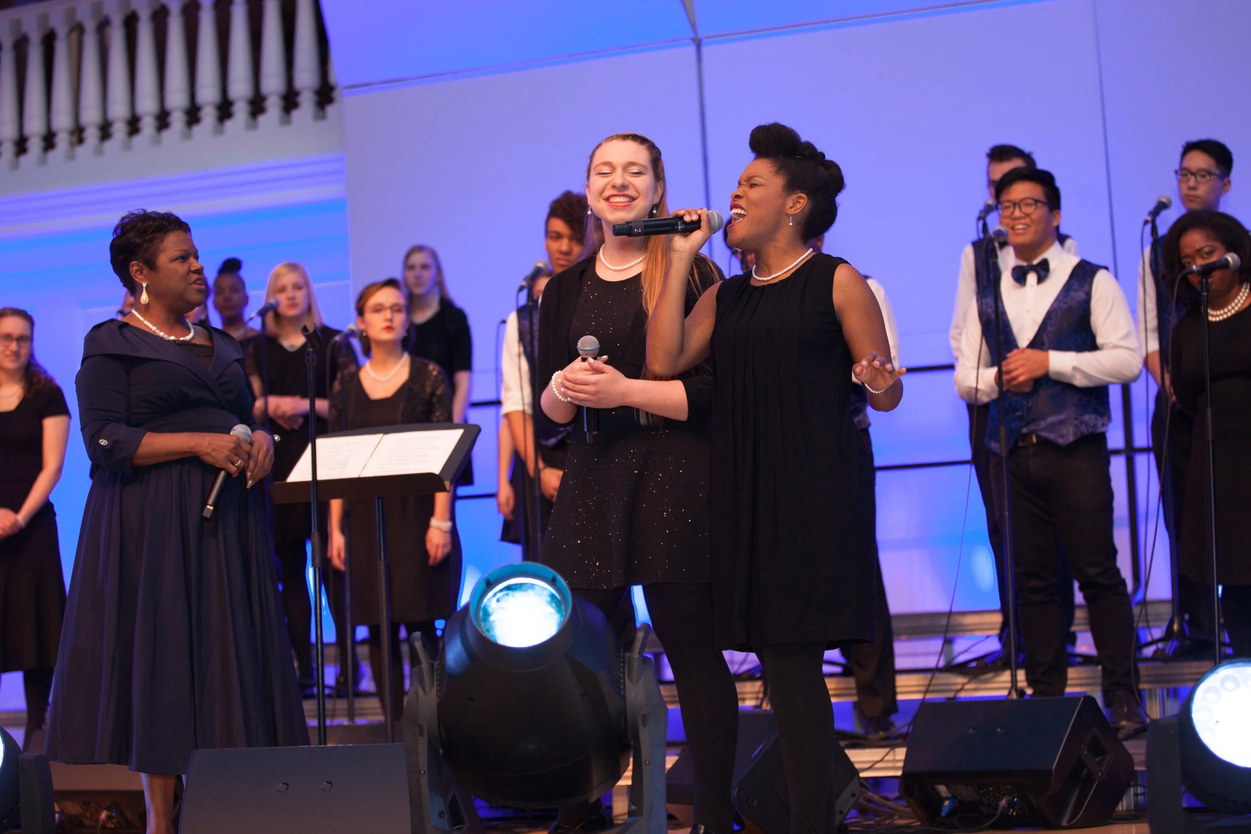 Group of choir singers on stage, two women in the front singing into microphones, others in the background, blue stage lighting, some women wearing black dresses with pearls, and some men in white shirts with vests.