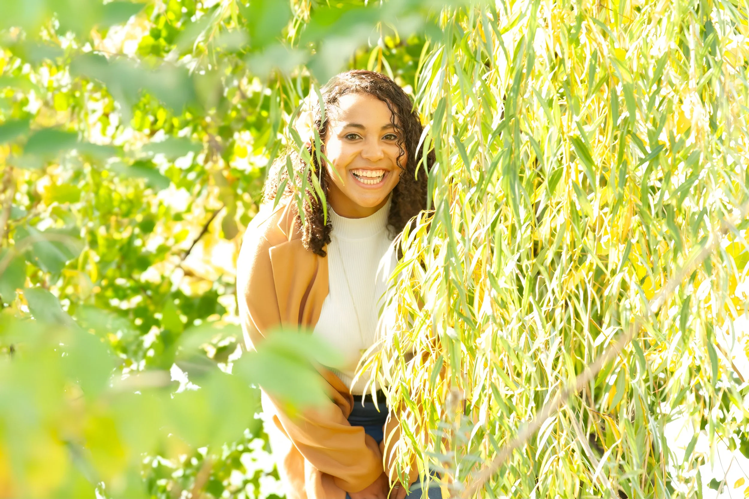 A woman smiling and enjoying the outdoors among hanging green and yellow leaves on a sunny day.