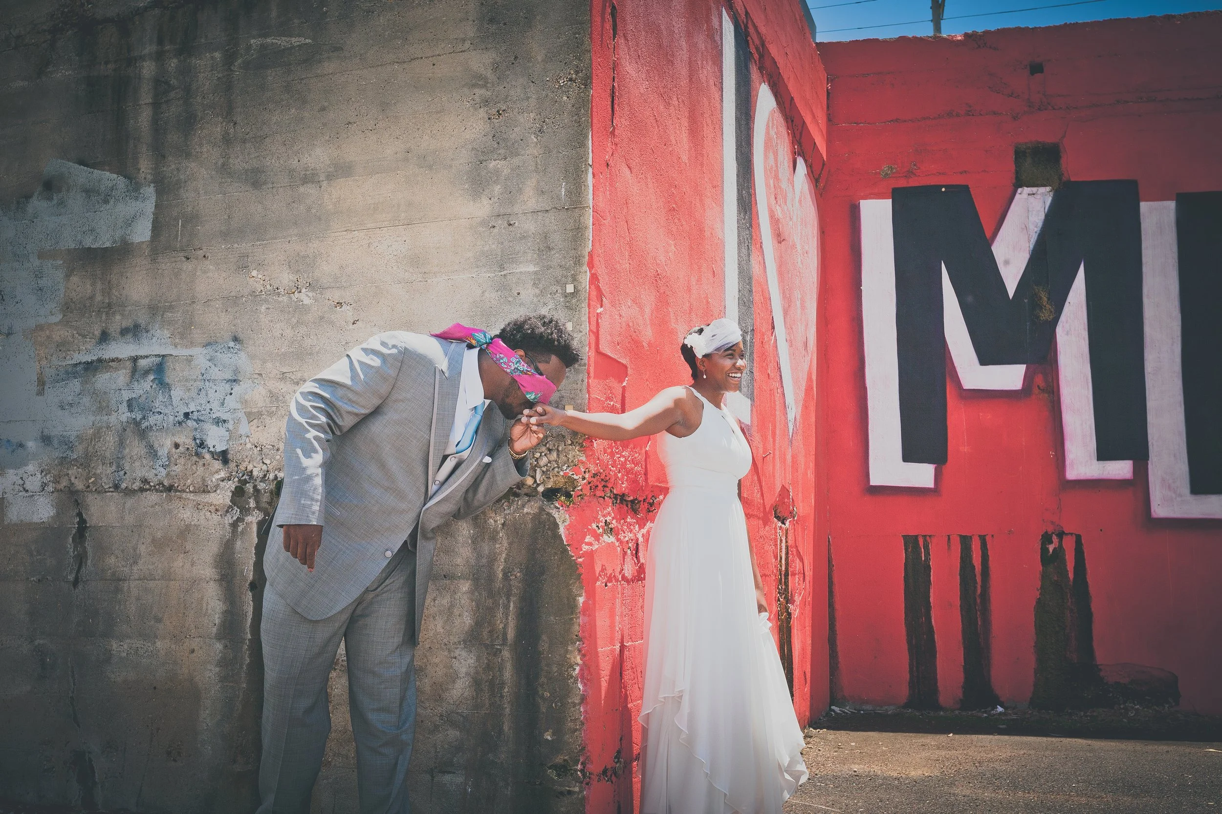 A woman in a white dress laughing while holding hands with a man in a gray suit with a pink scarf over his eyes, against a graffiti-covered wall.