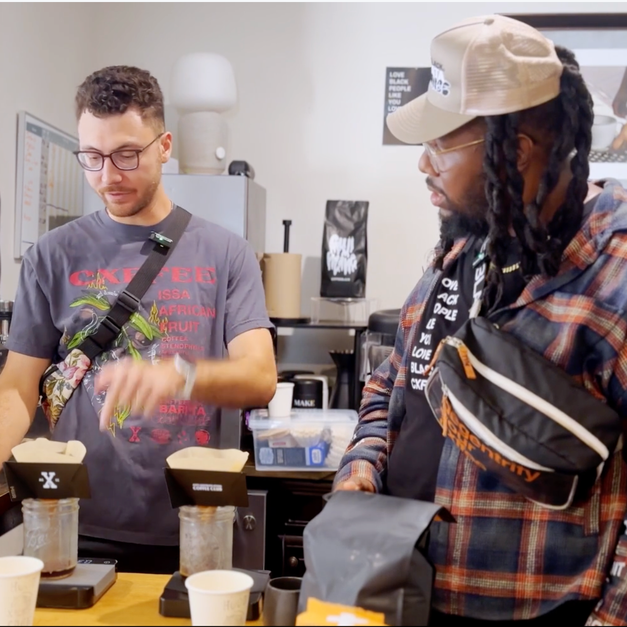 Two people in a coffee shop, one displaying bags of coffee beans and the other looking at the bags while wearing a beige cap and black crossbody bag.