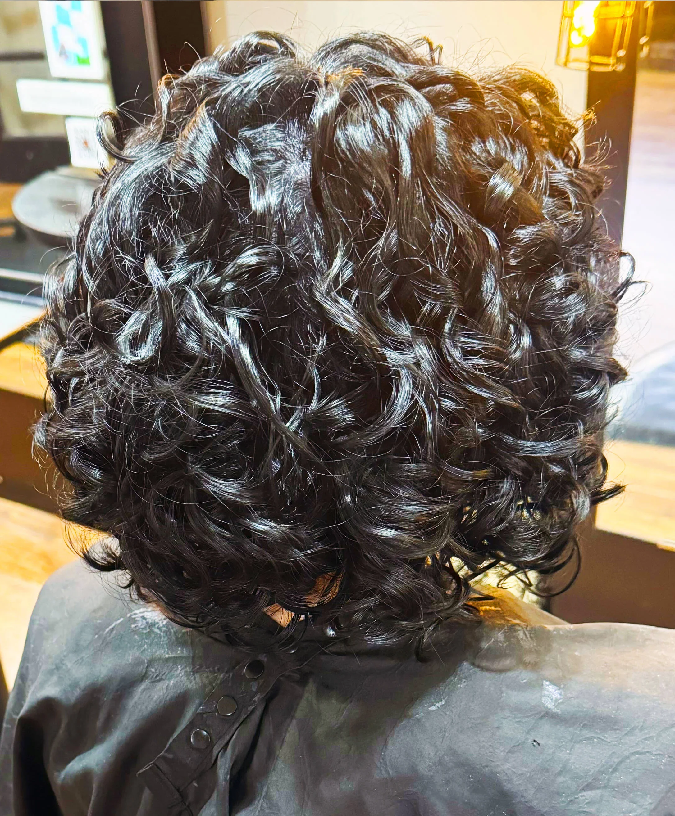 Back view of a person's head with short, black, curly hair styled in tight, defined curls.