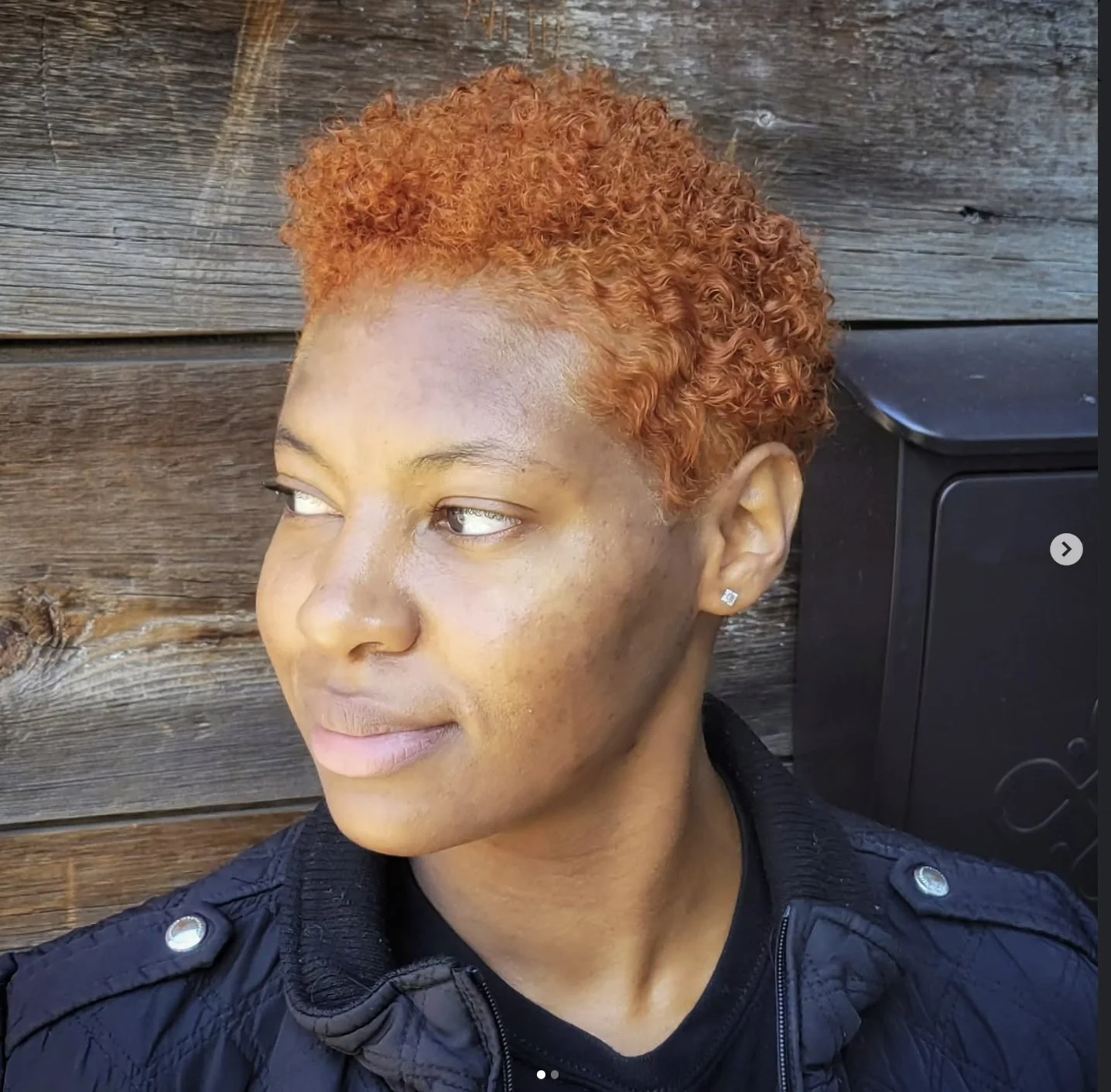 Close-up of a woman with short, curly, orange hair, wearing a black jacket with silver buttons, standing in front of a wooden wall.