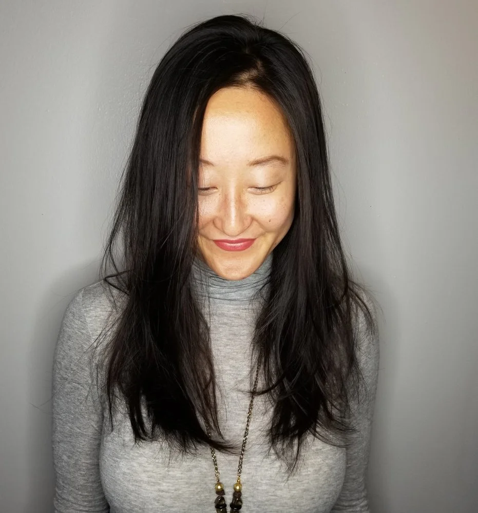 A woman with long black hair, wearing a gray turtleneck and a beaded necklace, has her eyes closed and is smiling softly against a plain gray wall.