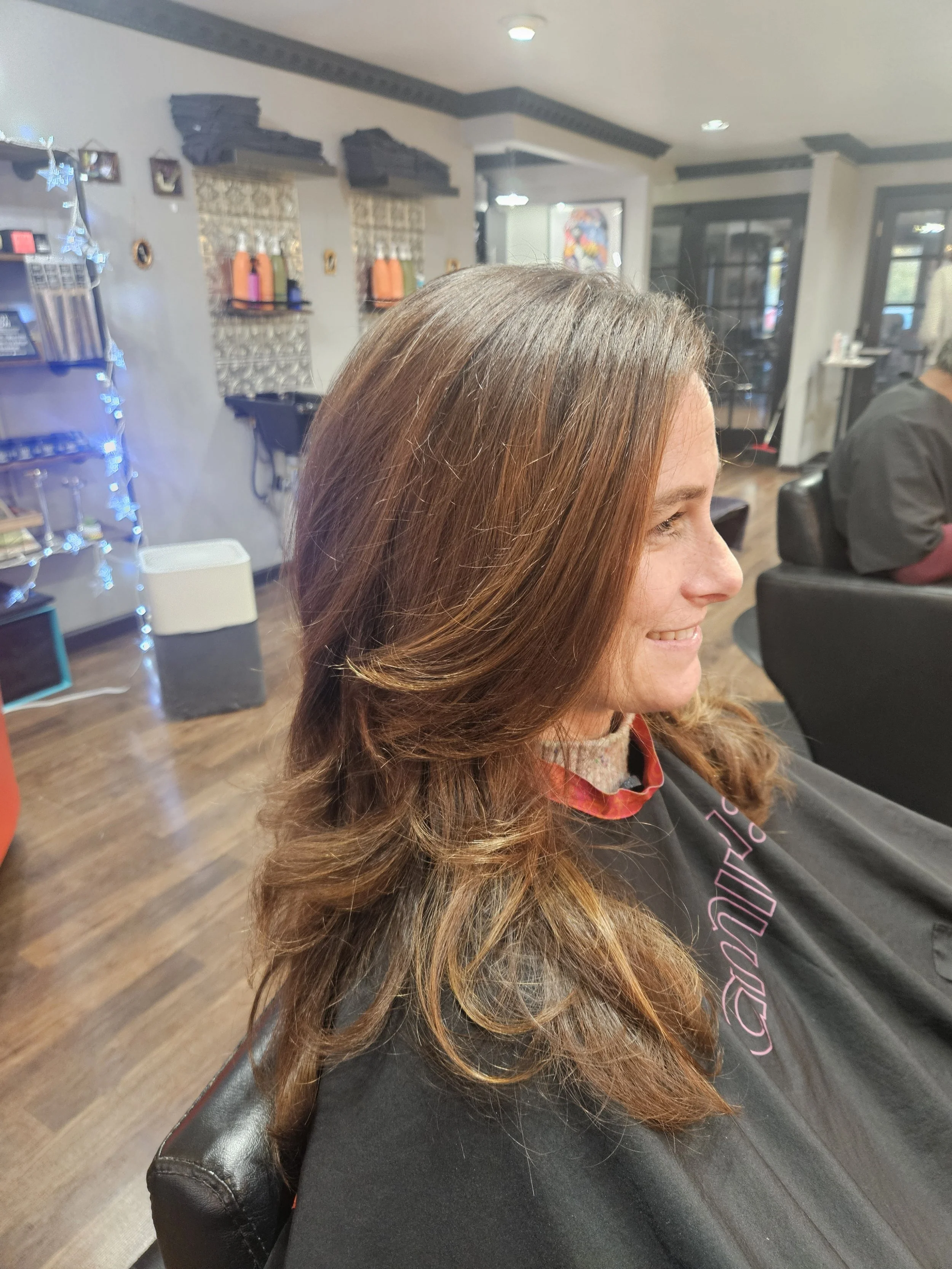 Side view of a smiling woman with long, wavy, brown hair sitting in a hair salon.