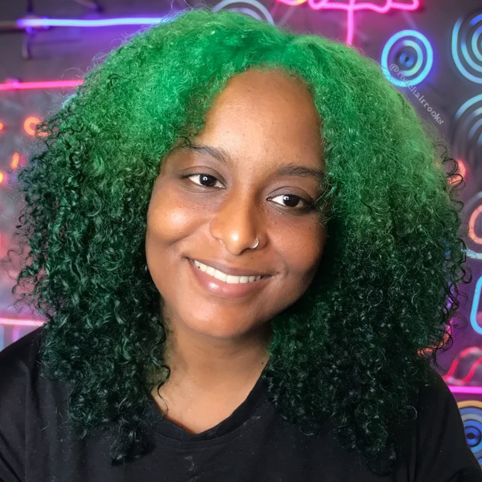 A woman with a bright smile, black curly hair with bright green dye at the top, and a nose ring. She is wearing a black shirt, and the background features colorful neon lights and abstract circular designs.