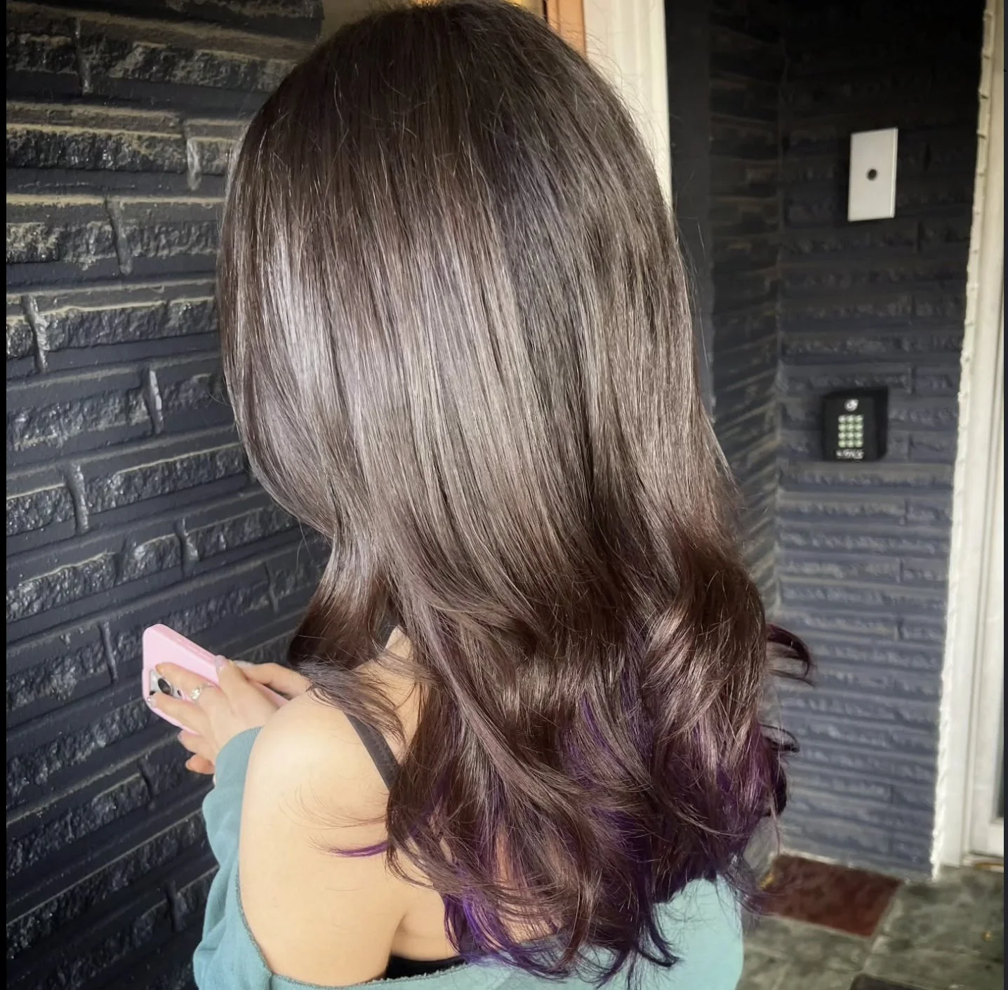 A woman with long, wavy hair with purple tips, looking at her phone, standing in front of a black brick wall with a white door and a keypad lock.