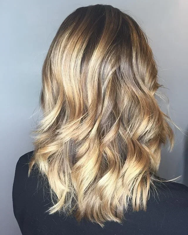 Back view of a woman with shoulder-length, wavy blonde hair with darker roots.