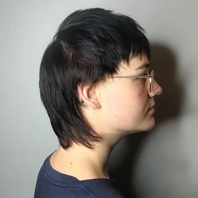 Profile of a woman with short, layered dark hair and glasses, facing to the right, against a plain wall.