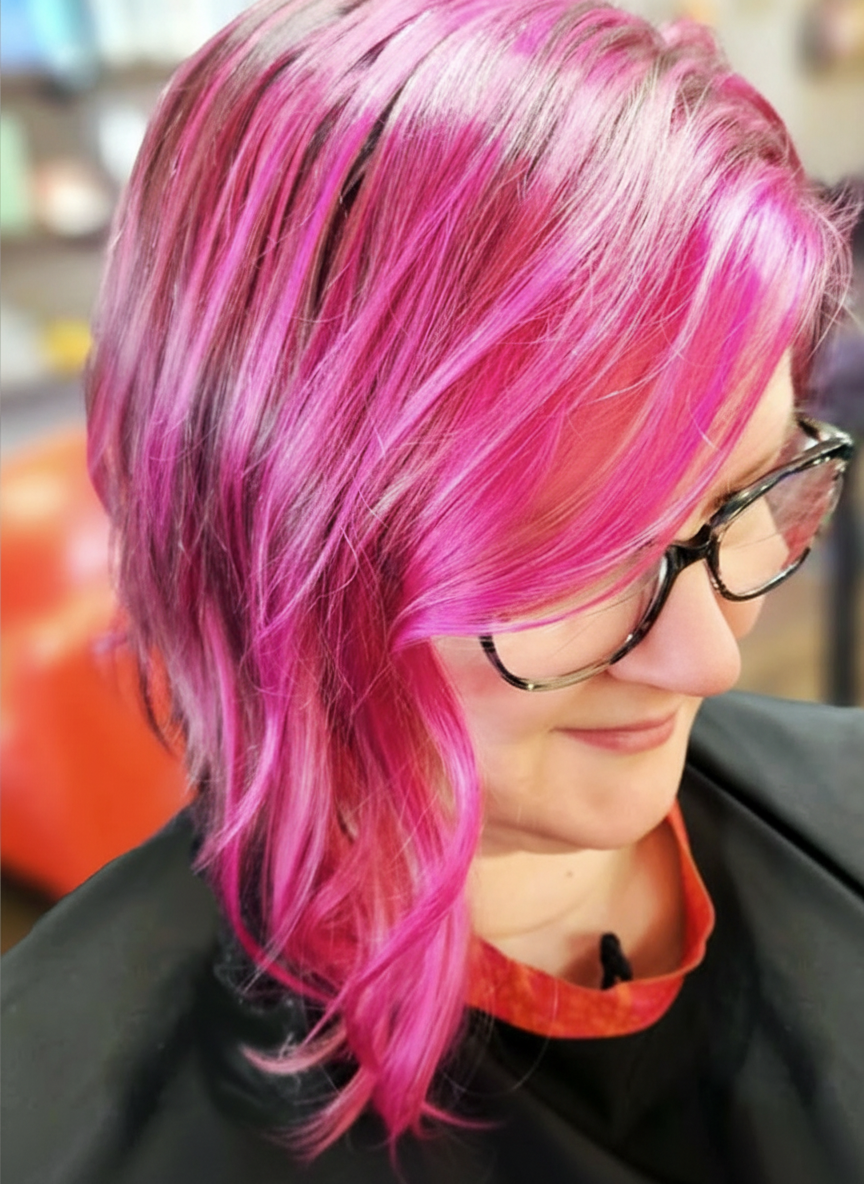 Fashionable woman with shoulder-length pink hair styled in loose waves, wearing glasses and a black salon cape in a hair salon.
