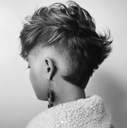 Black and white photo of a person with a modern, edgy hairstyle featuring shaved sides and longer, textured hair on top, wearing a fluffy sweater and an earring.