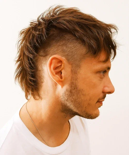 Side profile of a man with a mohawk hairstyle and a beard, wearing a white shirt and a silver necklace.