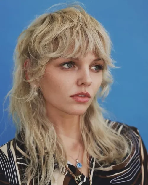 A woman with long, wavy blonde hair and blue eyes, wearing a black and white striped jacket, standing against a blue background.