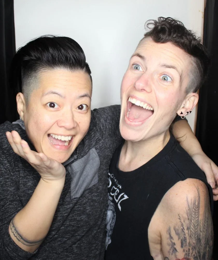 Two people smiling and celebrating, one with short black hair and the other with curly hair and tattoos on their arm.