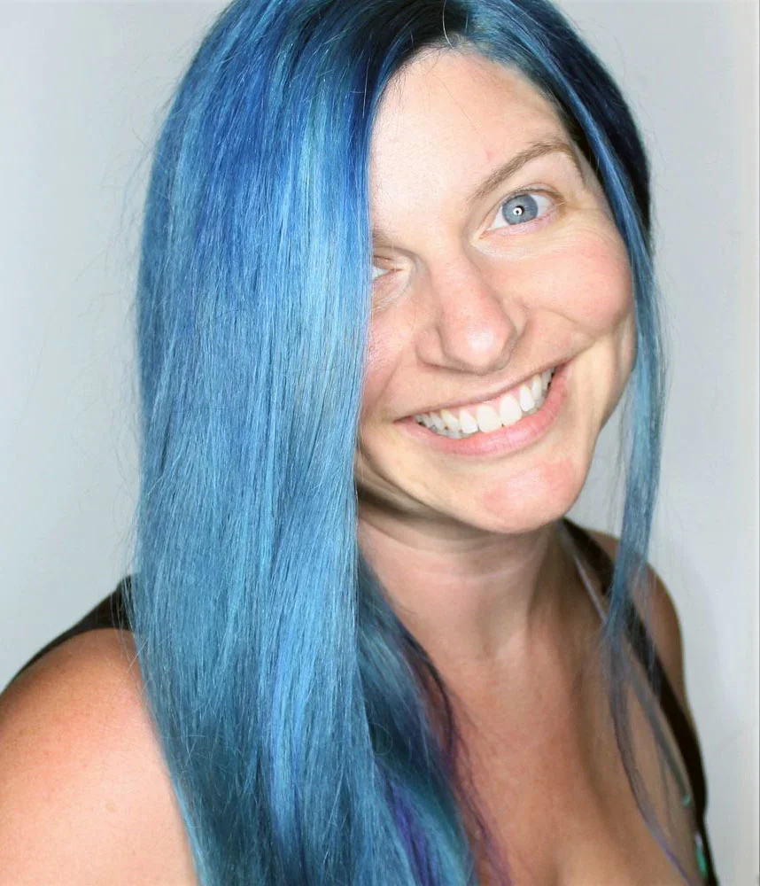 Close-up of a smiling woman with long, vibrant blue hair and fair skin, wearing a black top.