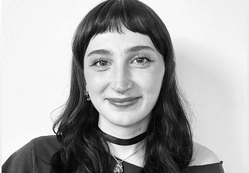 Black and white portrait of a young woman with dark hair, wearing a choker, hoop earrings, and a necklace, smiling slightly against a plain background.