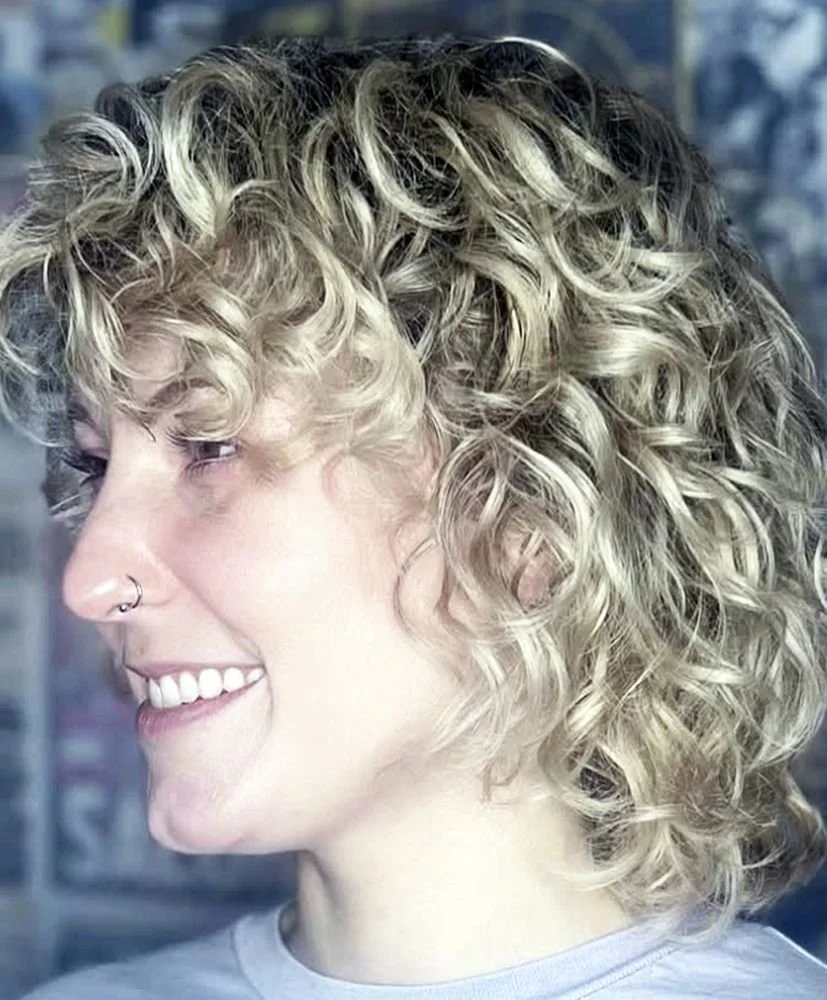 Side profile of a woman with short, blonde, tightly curled hair, wearing a nose ring and smiling.