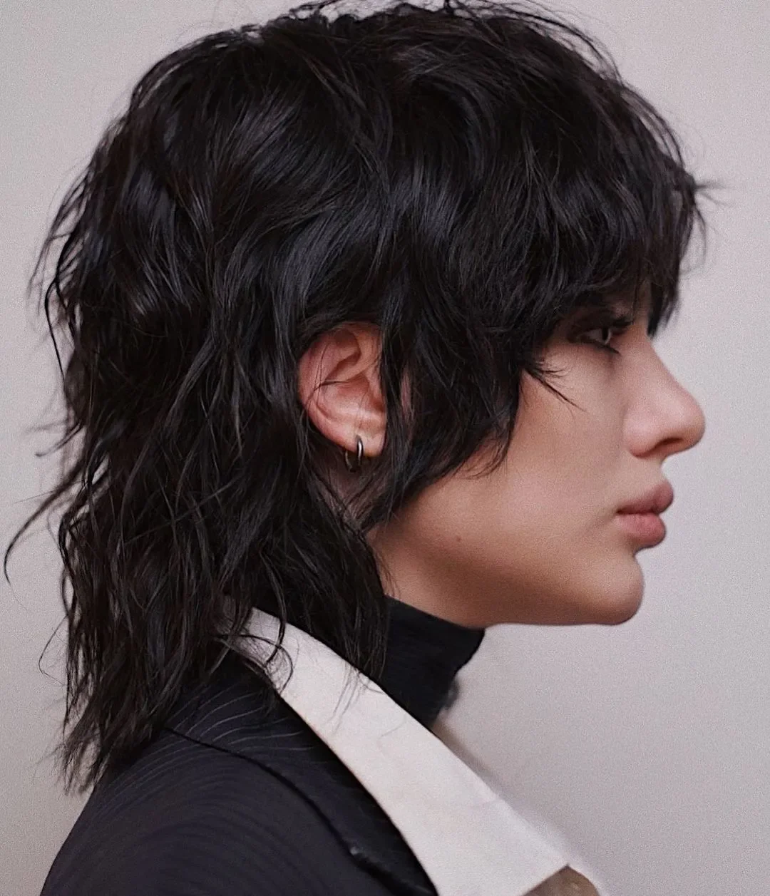 Side profile of a woman with wavy black hair, wearing a black turtleneck, blazer, and small hoop earrings.