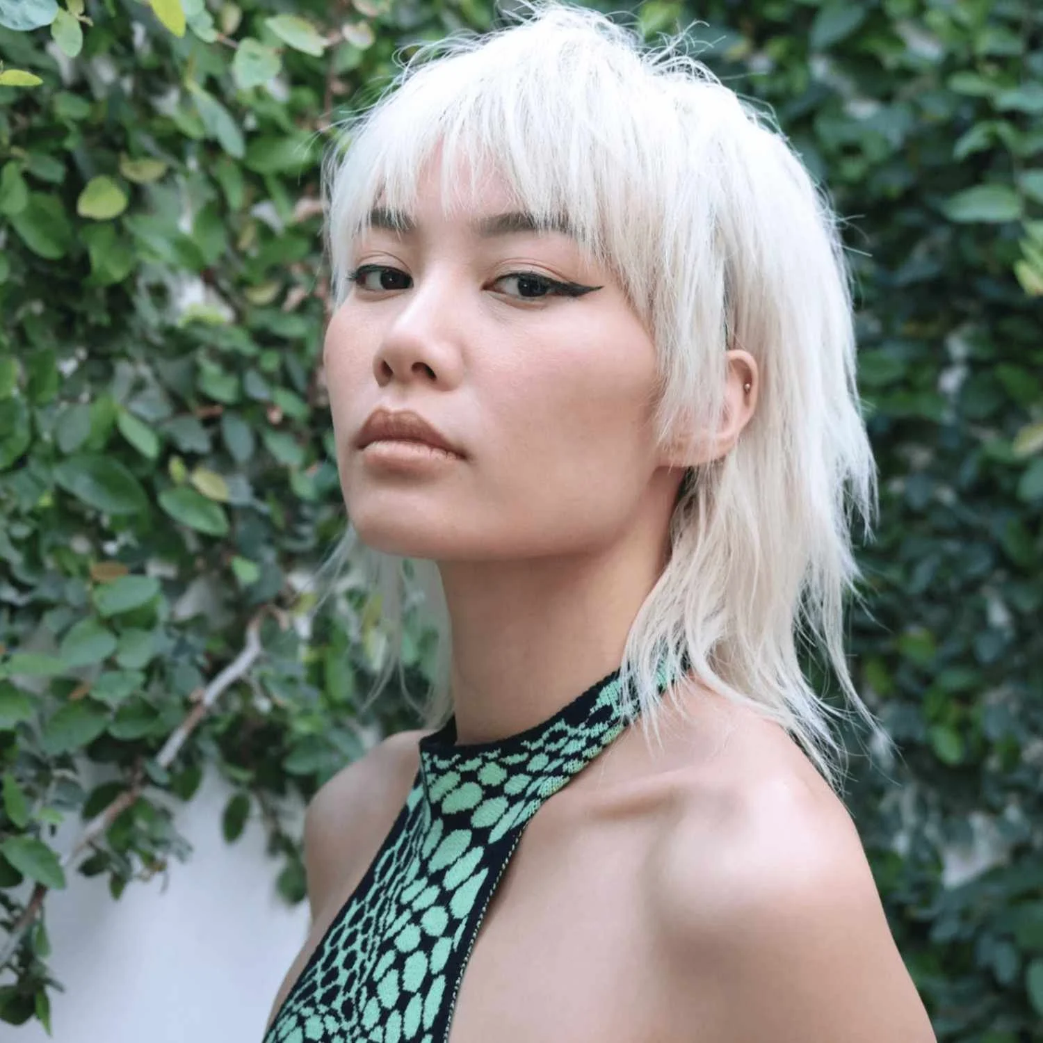 A woman with platinum blonde hair wavy shoulder-length hair, wearing a halter top with a black and turquoise pattern, standing outdoors in front of green leafy bushes.