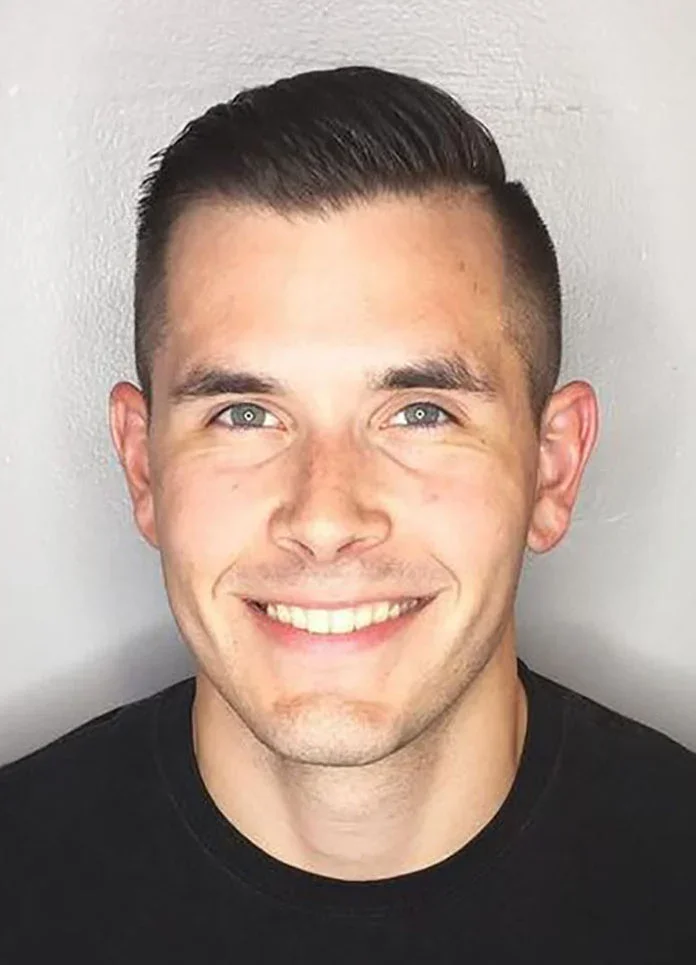 A young man with short, neatly styled dark hair, blue eyes, and a bright smile showing white teeth. He is wearing a black shirt and is photographed against a plain light-colored background.