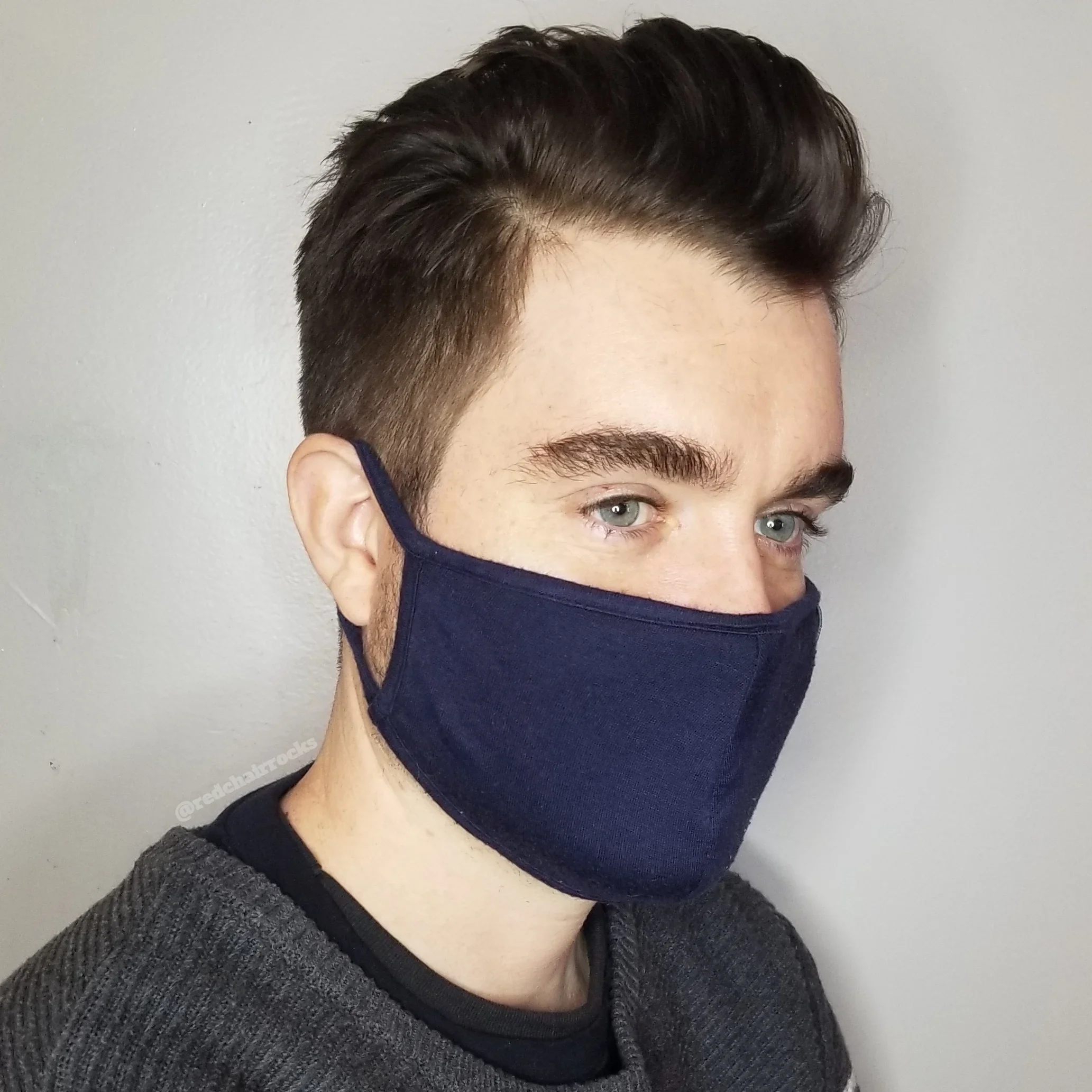 A young man with dark hair and blue eyes wearing a navy blue face mask and a gray knit sweater, standing against a plain wall.