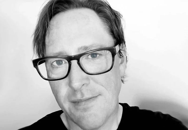 A black and white close-up portrait of a man with glasses and medium-length hair, smiling slightly, against a plain background.