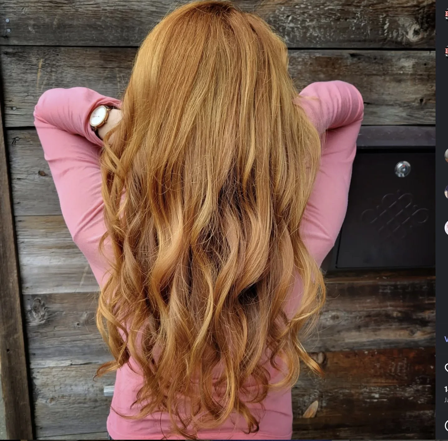 Person with long, wavy, reddish-blonde hair wearing a pink long-sleeved top and a wristwatch, standing with back to camera in front of weathered wooden wall.