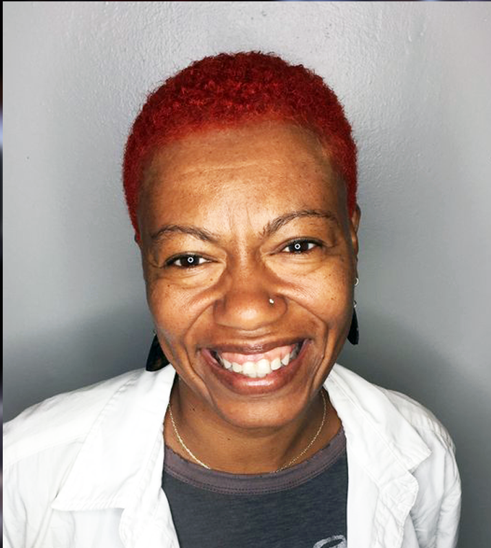 A woman with short red hair, smiling, wearing a white shirt over a gray top, with earrings, a nose piercing, and a necklace, against a light gray background.