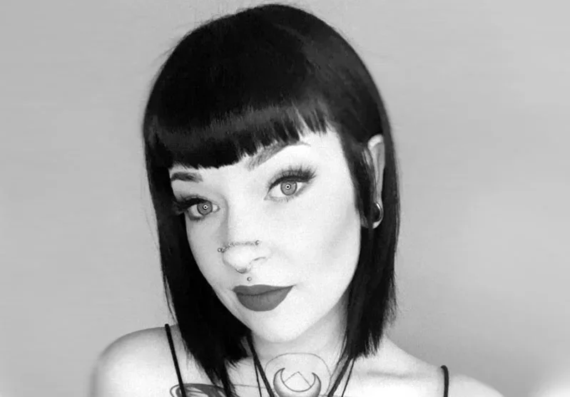 A woman with black hair, heavy eye makeup, dark lipstick, and facial piercings, wearing a black spaghetti strap top, in front of a plain wall.