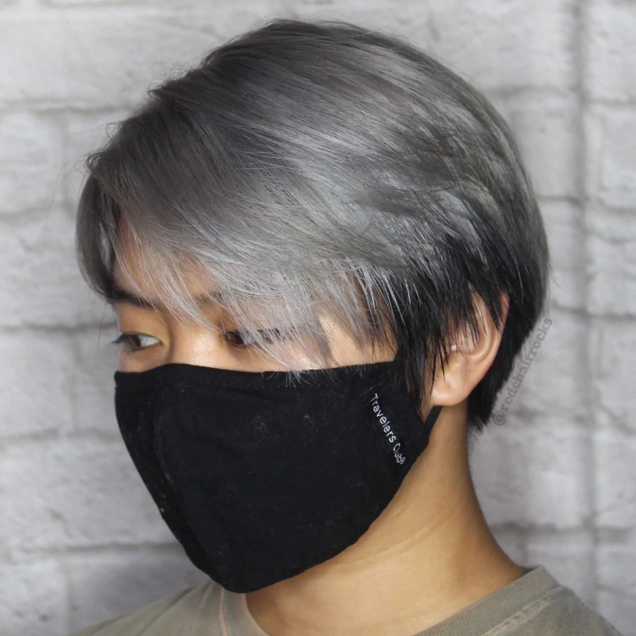 Person with short, layered hair dyed silver and black, wearing a black face mask, standing against a white brick wall.