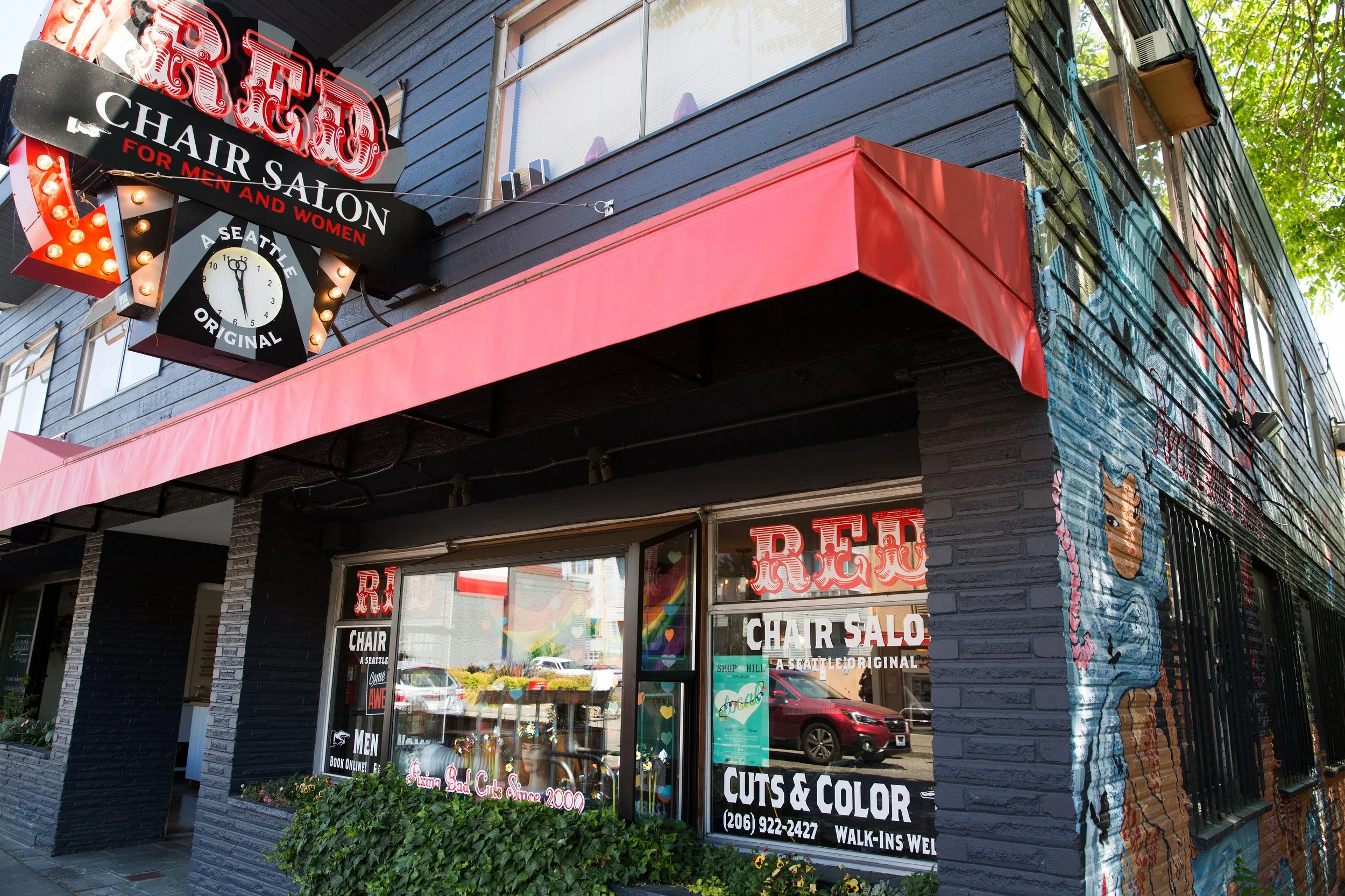 Exterior of a hair salon called 'Red Chair Salon' with red and black signage, large front window displaying salon services, and a mural of a woman with blue hair on the building's corner.