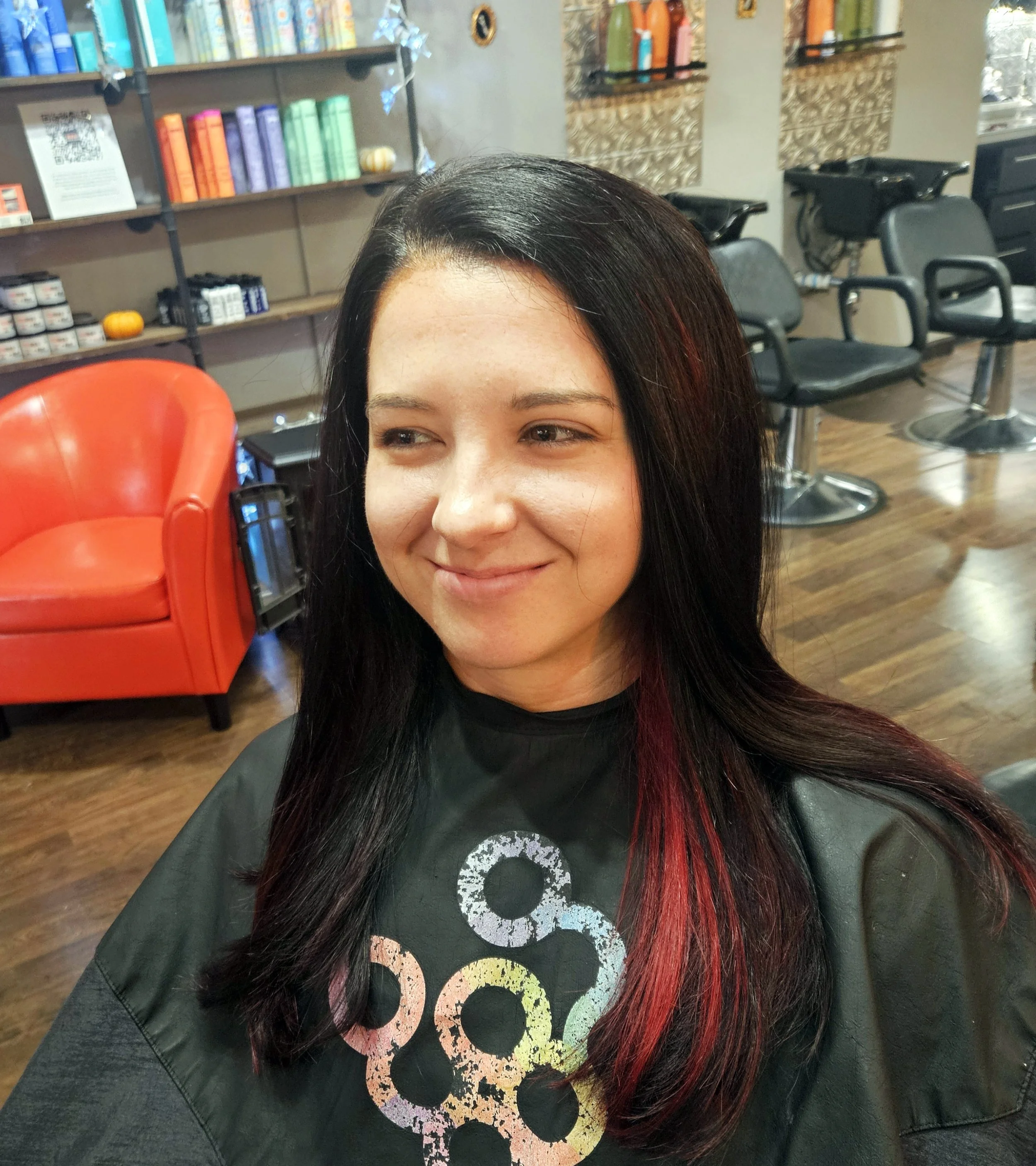 A woman with long black hair and red highlights smiling in a salon chair, wearing a cape with colorful circles, with salon chairs, shelves with hair products, and decorative items in the background.