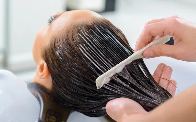 Person receiving a hair treatment at a salon, applying conditioner or treatment to long, dark hair with a small comb.