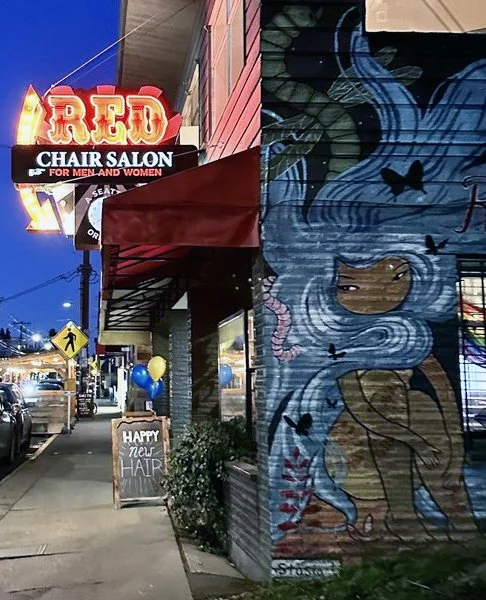 Street scene at dusk showing a mural of a girl with long flowing blue hair with stars and veiled in pink on a brick wall, neon sign for "Red Chair Salon" for men and women, and a chalkboard sign that reads "Happy New Hair" with balloons tied to a plant outside the salon.