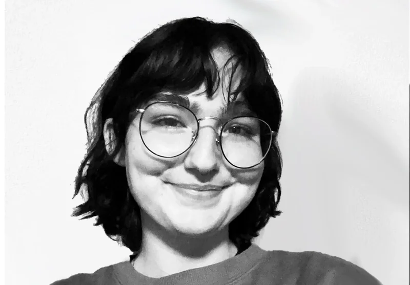Black-and-white photo of a woman with wavy dark hair, wearing glasses, smiling, against a plain background.