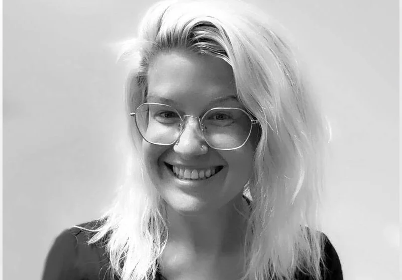 A woman with long, wavy blond hair, wearing glasses, smiling, against a plain background.