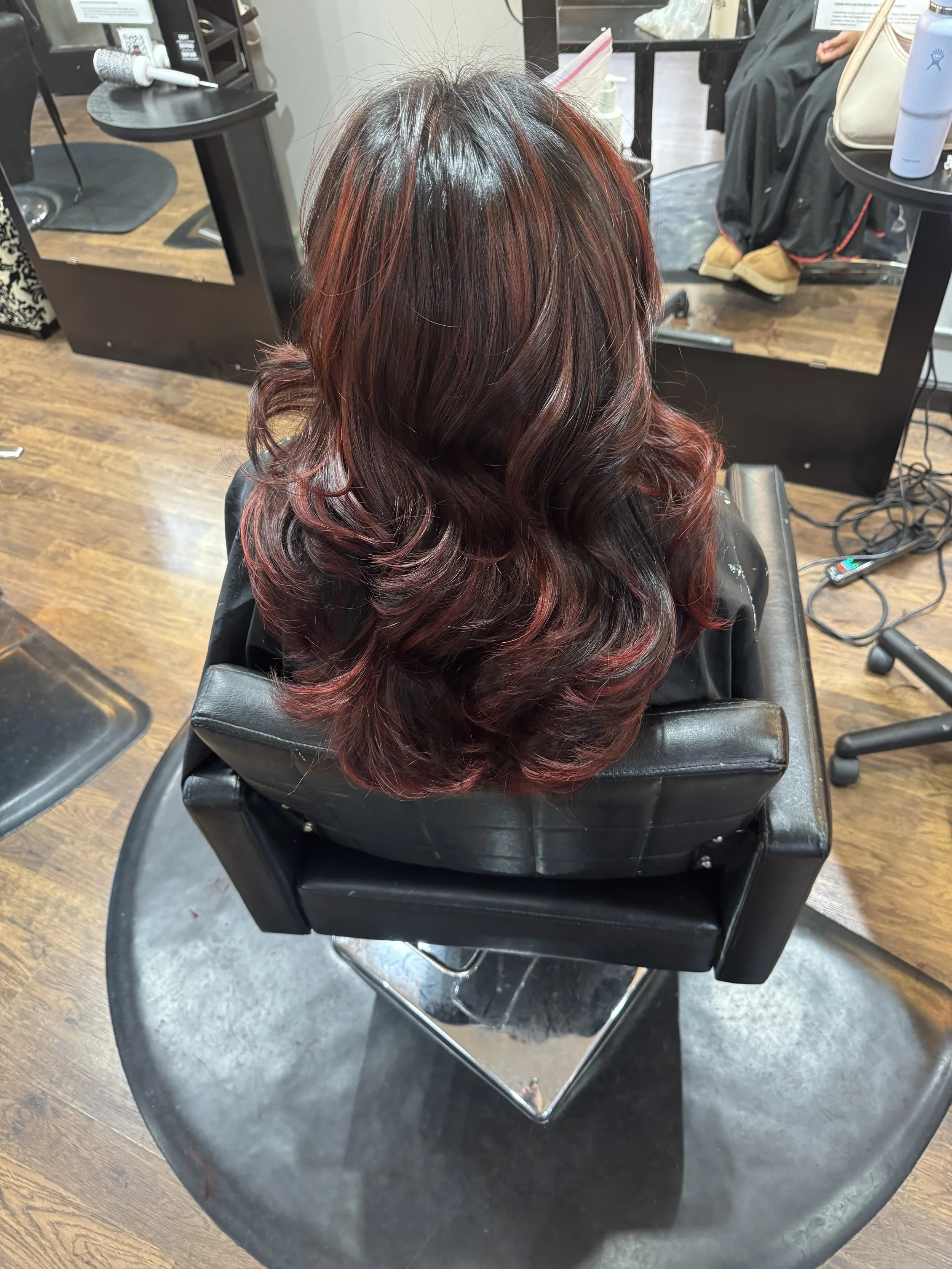 Back view of a woman with shoulder-length wavy hair dyed in shades of red and burgundy, sitting in a salon chair.