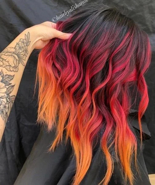 A woman with long wavy hair featuring a vibrant gradient from dark at the roots to bright orange at the ends, with pink highlights.